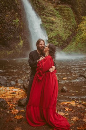 A couple shares an intimate moment by a cascading waterfall with red dress contrasting against autumn scenery.