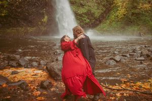 Two people in dramatic poses against a waterfall backdrop with autumn leaves on the ground.