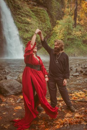 A dancing couple in autumn with one in a flowing red dress and the other in dark clothing near a waterfall.