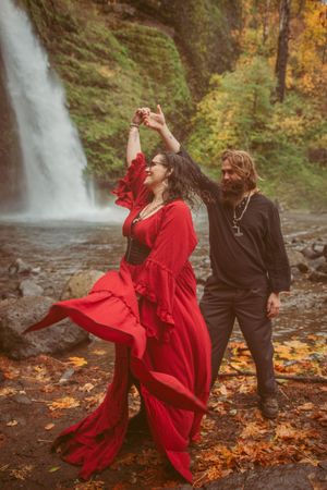 Dancers in red and black perform expressive movements against a forest waterfall backdrop with autumn foliage.
