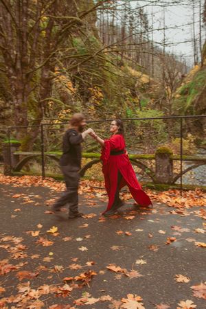 Two people posing dramatically on a walkway covered in autumn leaves with a forest backdrop.