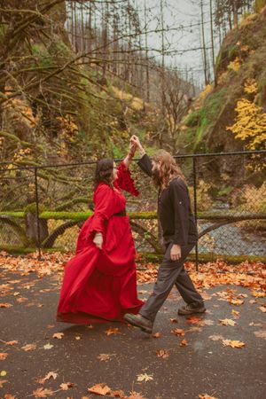 A couple dancing in autumn leaves on a path with one wearing a flowing red dress and the other in dark casual wear.