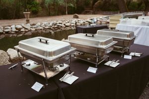 Row of stainless steel catering chafers lined up on black tablecloth for buffet service at an event.
