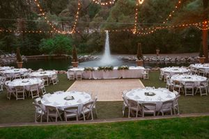 Outdoor wedding reception setup with round tables and string lights near a fountain.
