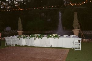An elegant outdoor reception dinner table setup with white linens and floral centerpieces near a fountain at dusk.