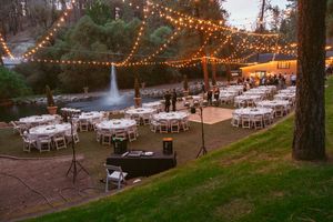 Outdoor wedding venue at dusk with string lights and white tables arranged under trees.