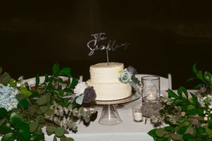 Simple white wedding cake with topper displayed among green foliage decorations.