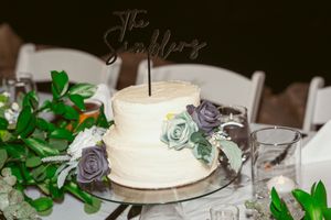 Elegant white wedding cake decorated with purple and sage sugar flowers on a classic cake stand.