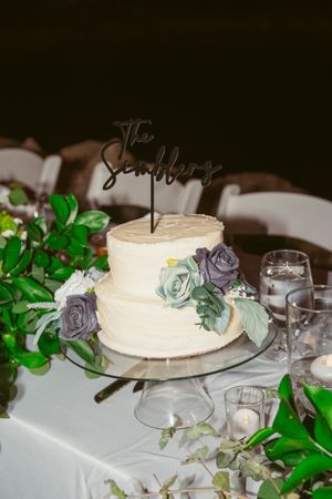 Two-tier white wedding cake decorated with blue and purple roses and a modern black cake topper.