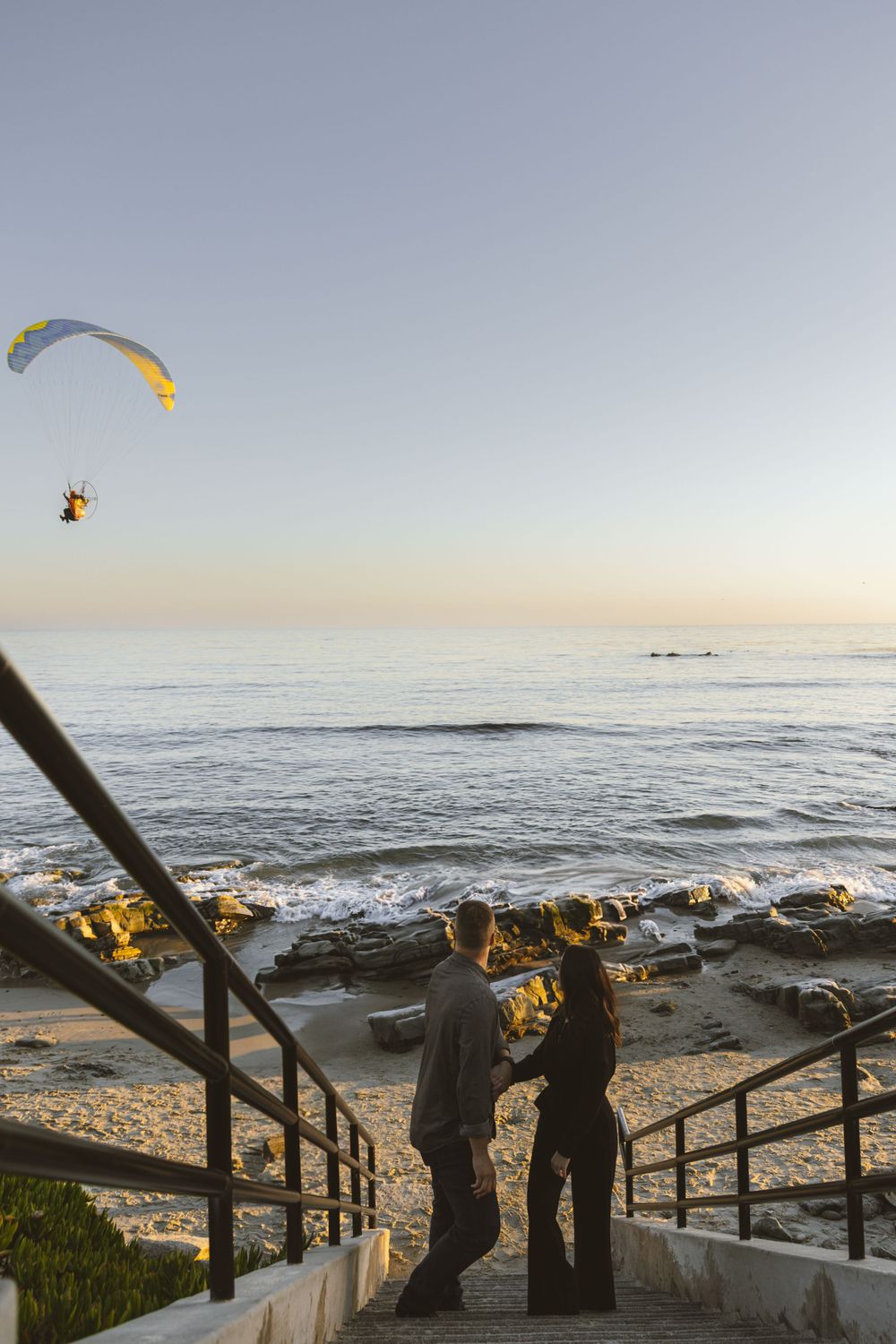 Couple embraces on beach stairs at sunset while paraglider soars in background.