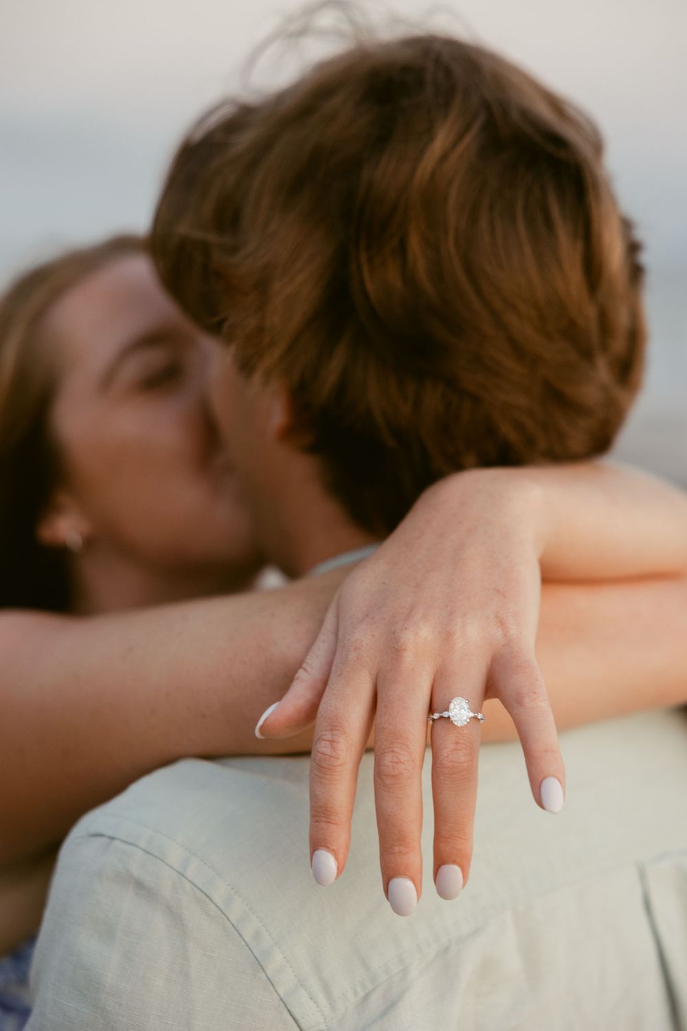 Couple sharing a kiss while showcasing a diamond engagement ring on her hand resting on his shoulder.
