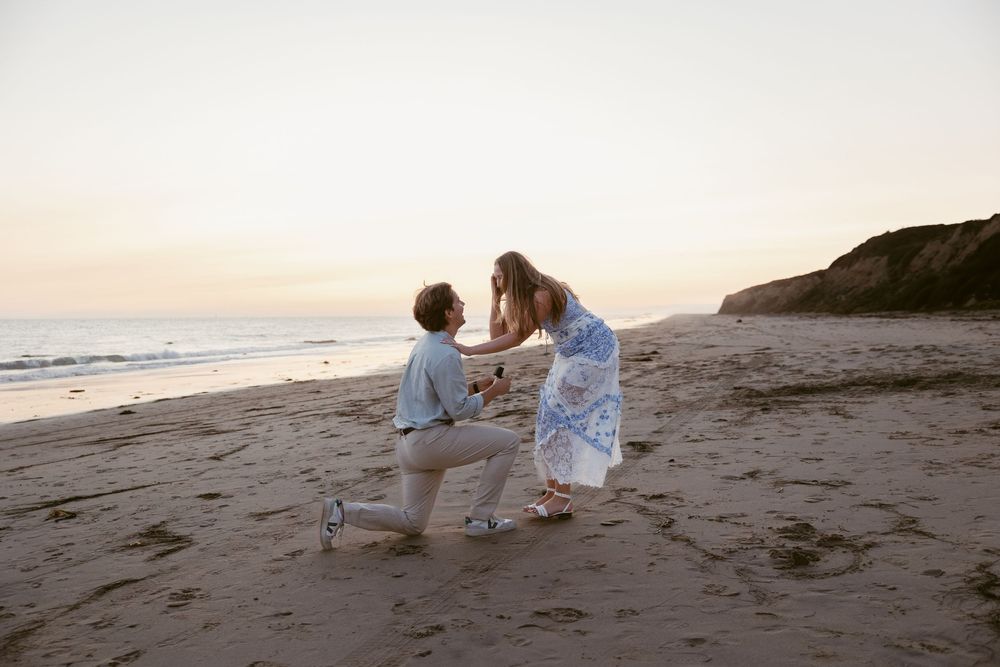 Man proposes to woman on a sandy beach at sunset, kneeling before her in a romantic coastal setting.