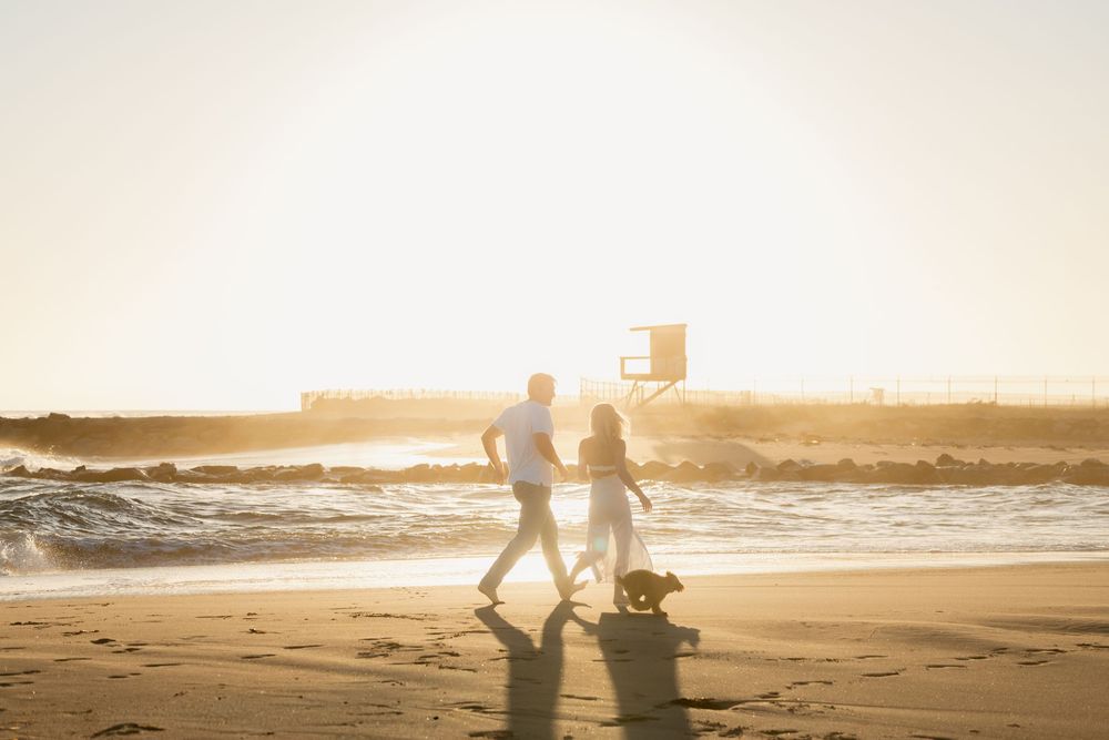 Couple walking dog on sunny beach at golden hour with waves and lifeguard tower in background.