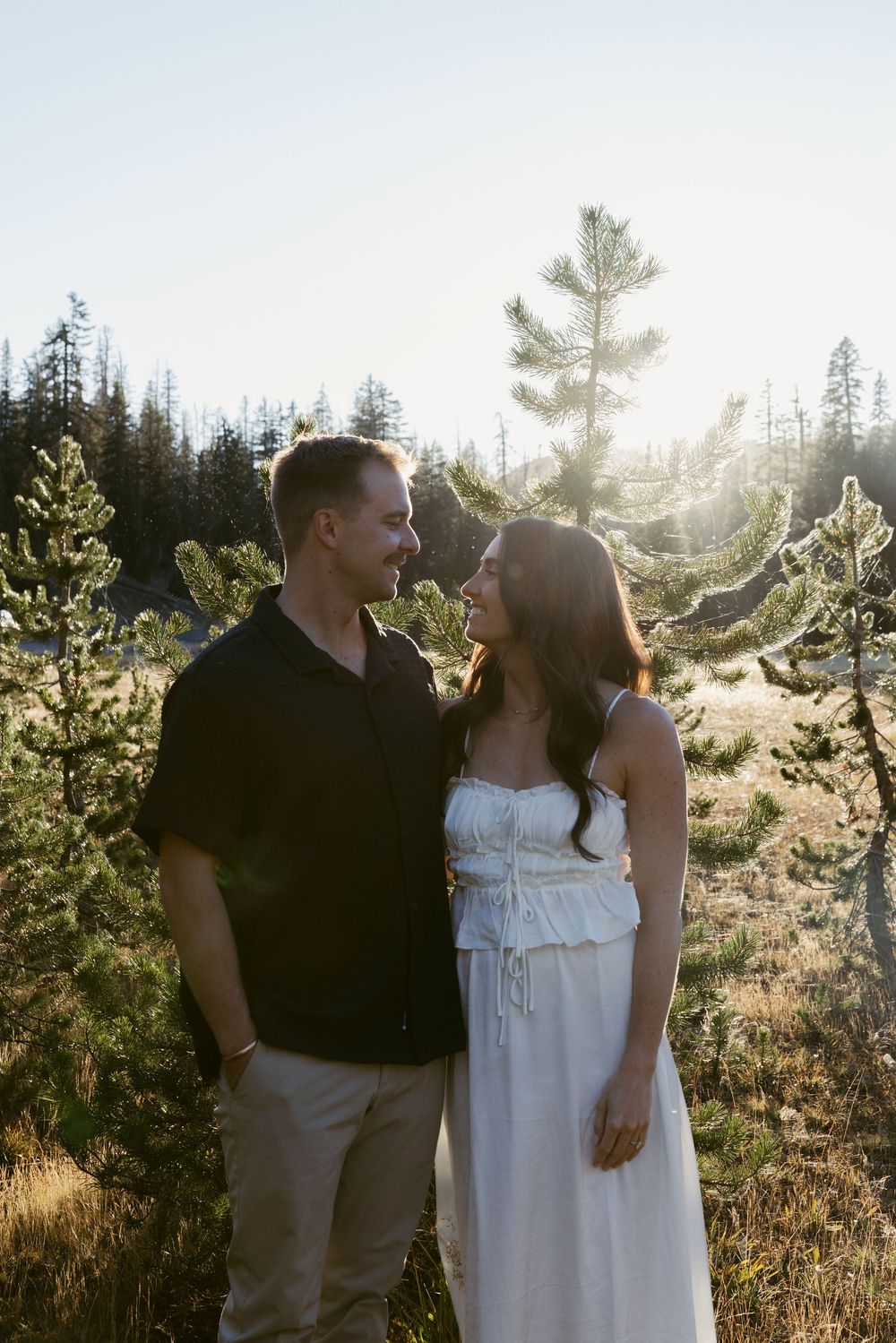 Couple smiling at each other in a sunlit forest clearing, man in black shirt and woman in white dress.