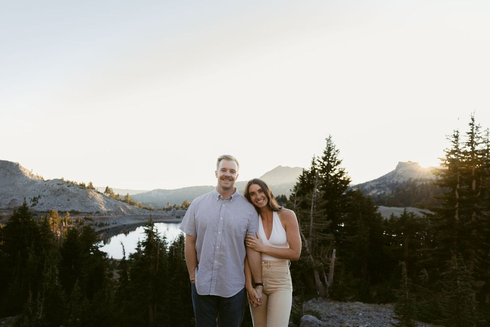 Couple smiling together at golden hour with mountain lake and pine trees in scenic backdrop.