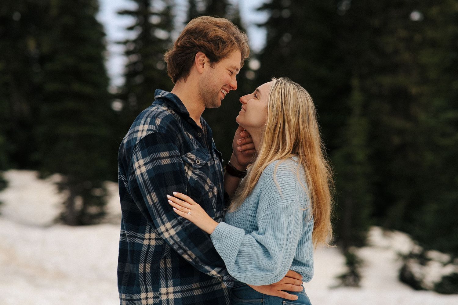 A couple embraces in a snowy forest setting while wearing winter clothing and plaid flannel.