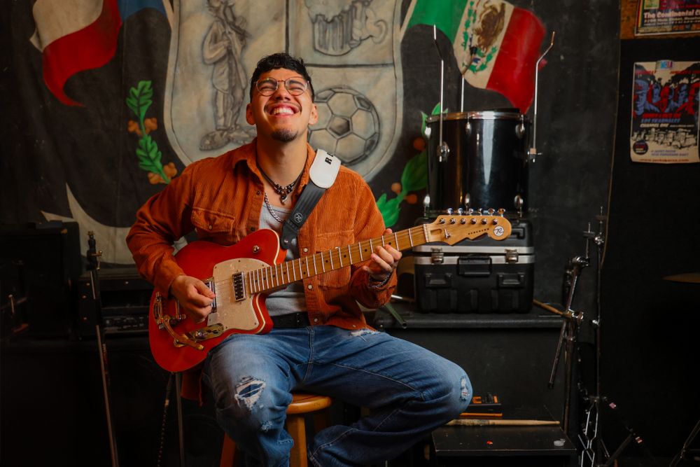 Musician in orange jacket plays guitar against dark wall with Mexican flags and decorative backdrop.