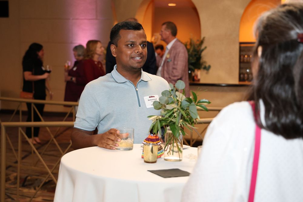 Man in light blue polo shirt socializing at a networking event, holding a drink near a white high-top table.