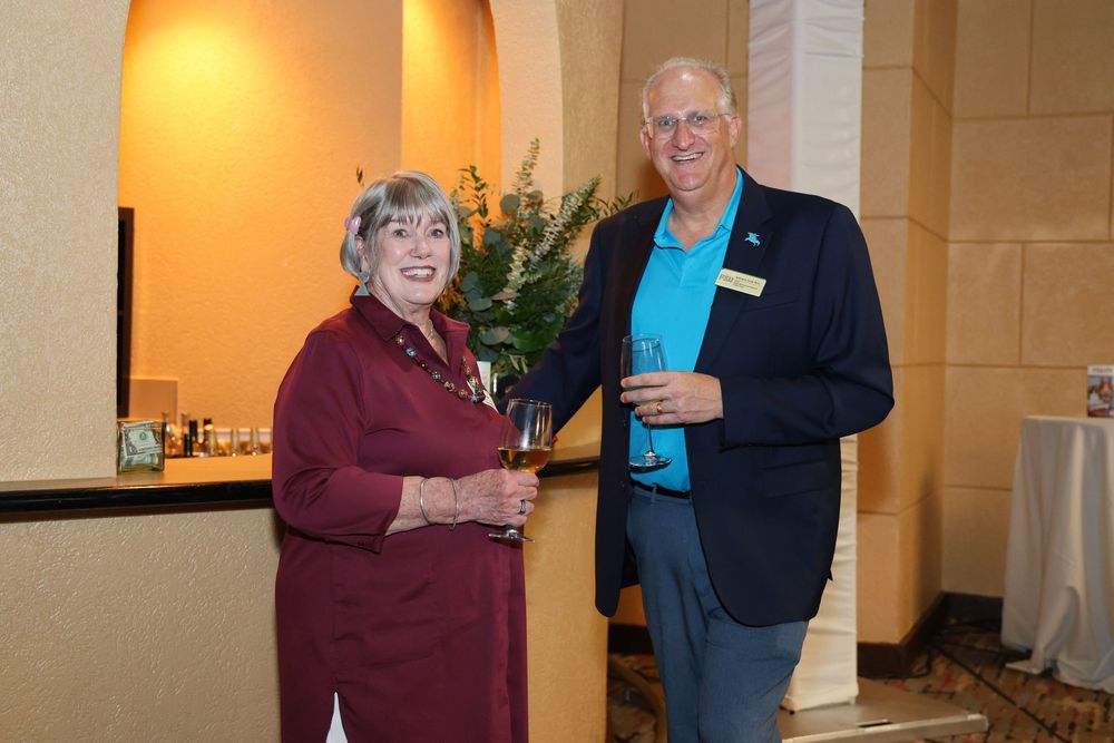 Two smiling adults holding wine glasses at an indoor reception event in an elegantly decorated venue.