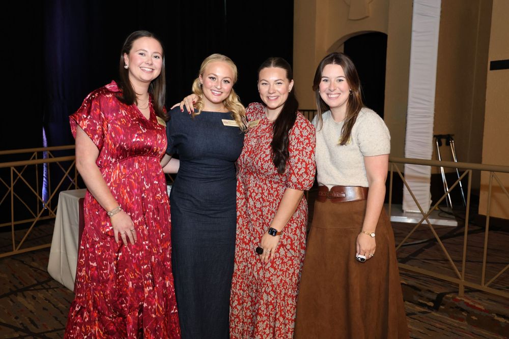 Four women smiling and posing together at a formal event in an elegant ballroom setting.