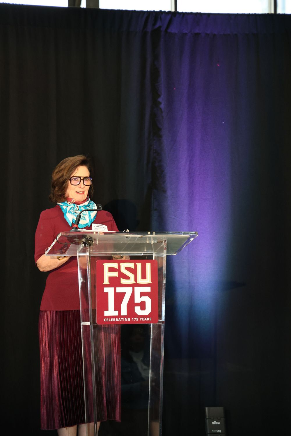 Woman in burgundy dress speaking at FSU 175th anniversary celebration podium at formal event.
