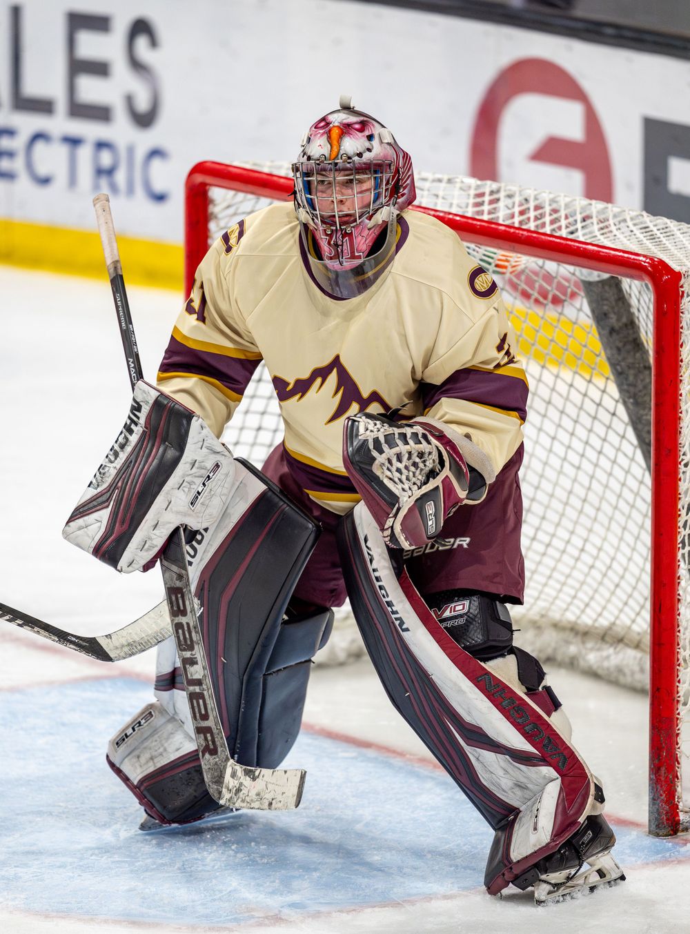 Hockey goalie in cream and maroon uniform stands ready in defensive position at net.