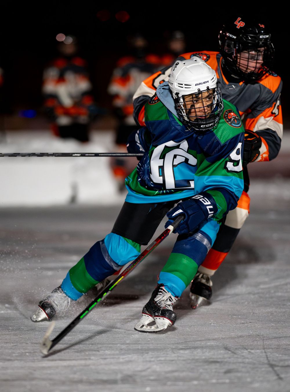 Youth hockey player in teal jersey battles opponent during nighttime game.