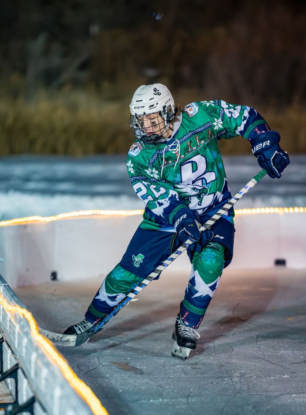 Hockey player in green and blue jersey skates near illuminated boards during evening match.