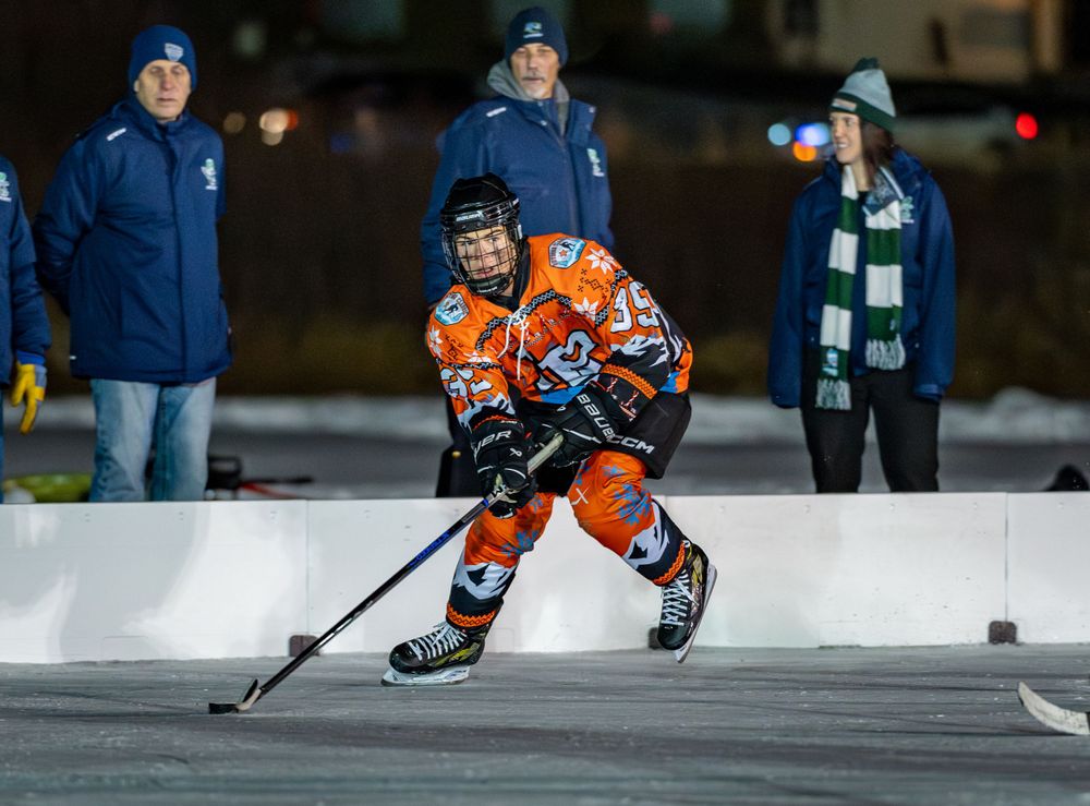 Hockey player in orange jersey controls puck during outdoor game with spectators watching.