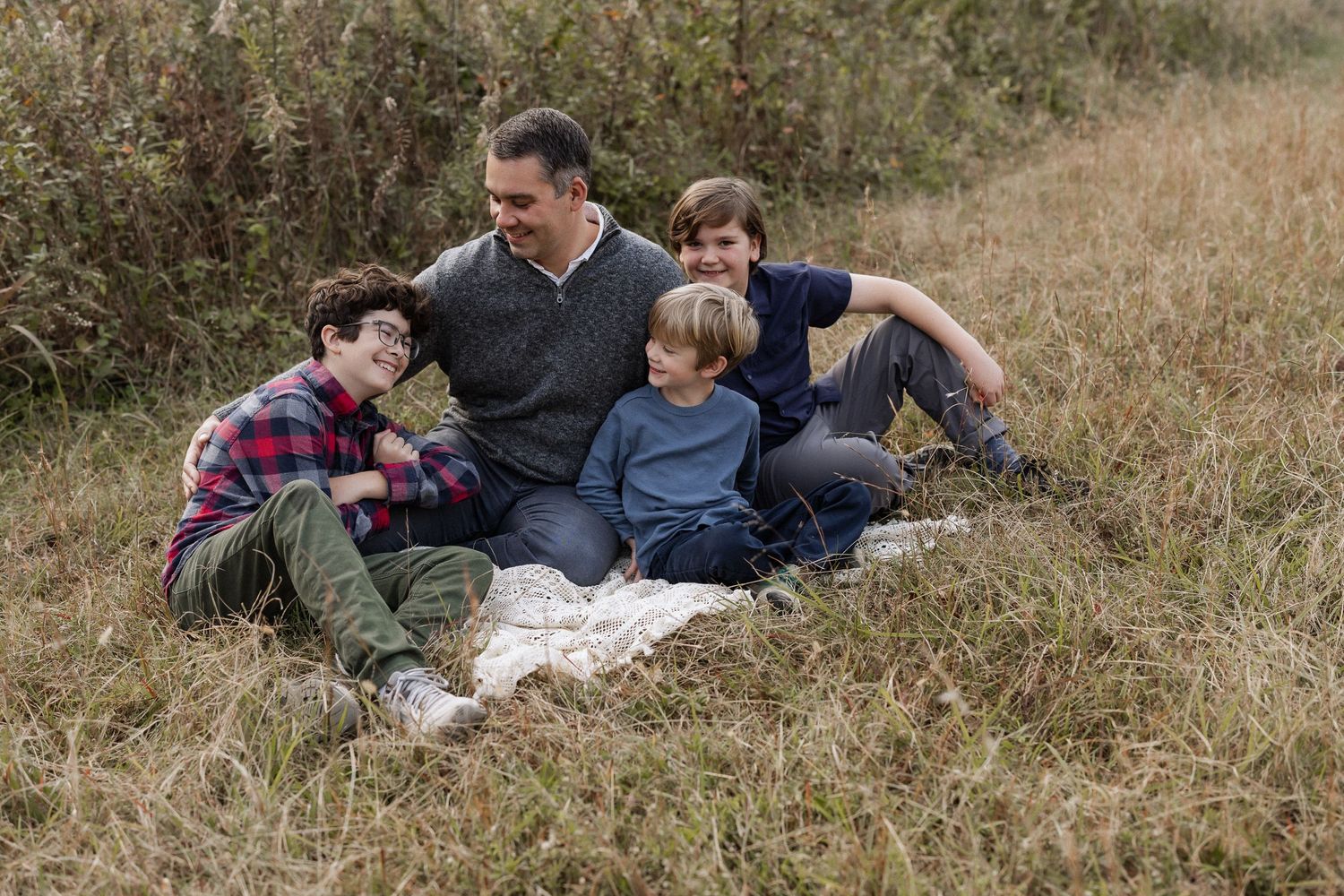 A family sits together on a blanket in tall brown grass during an outdoor autumn photo session.