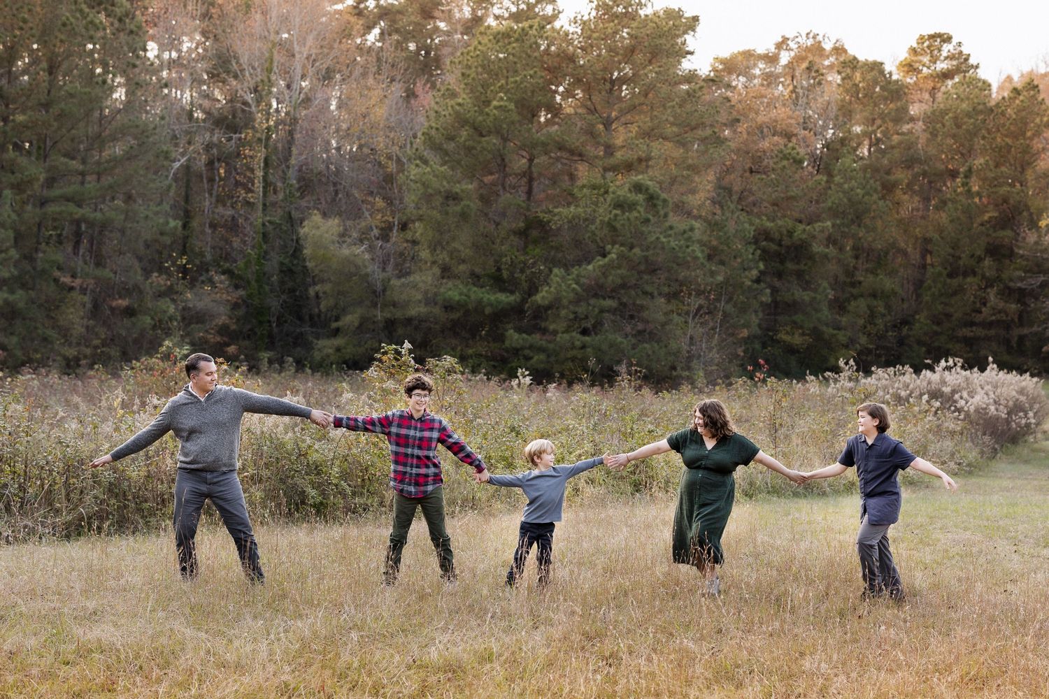 A group holds hands in a line while standing in a golden autumn field at Duke Homestead at sunset with pine trees in the background.