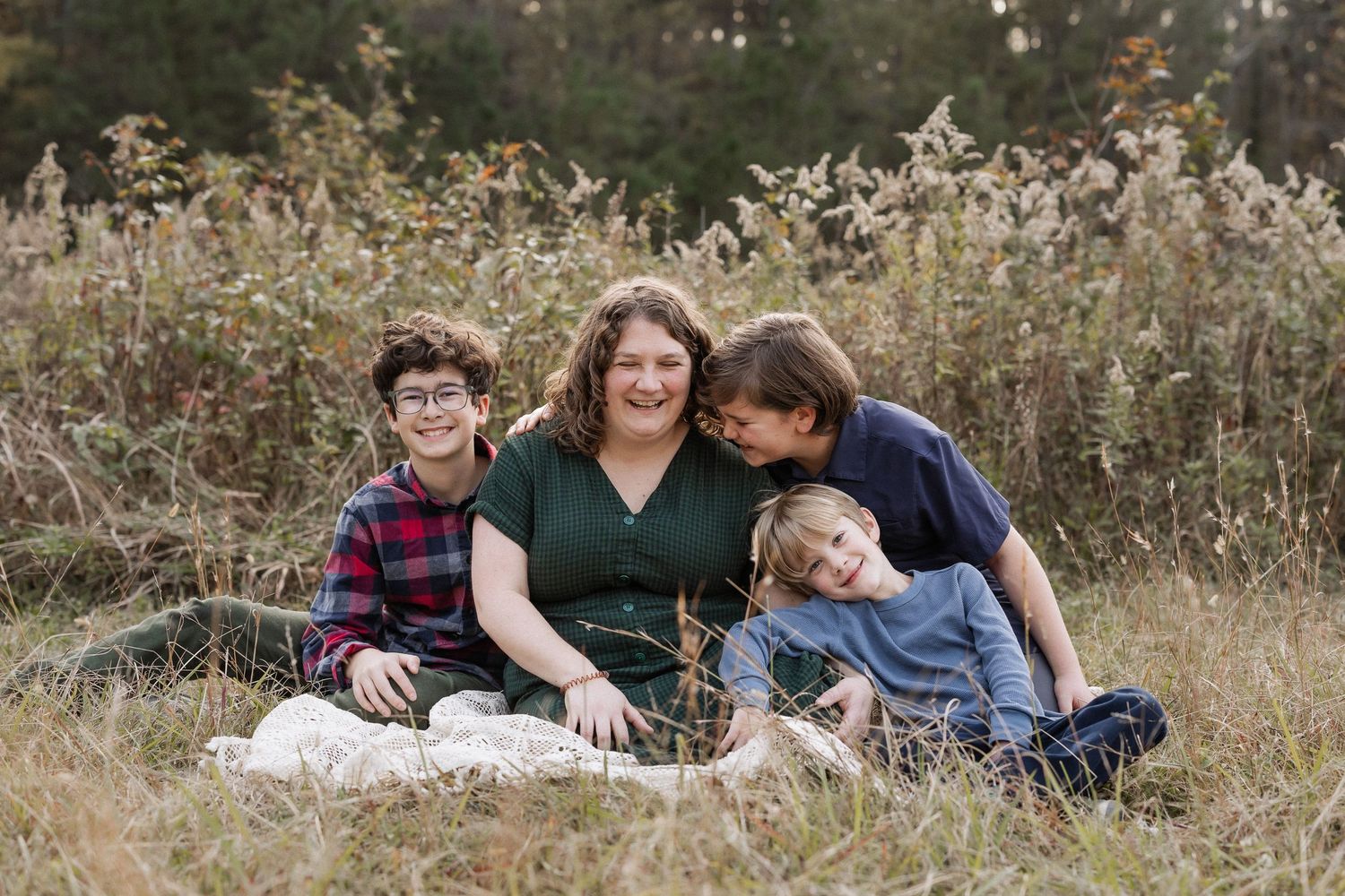 A family sits together on a blanket  in a field with tall grass and wildflowers at Duke Homestead during an outdoor photo session.