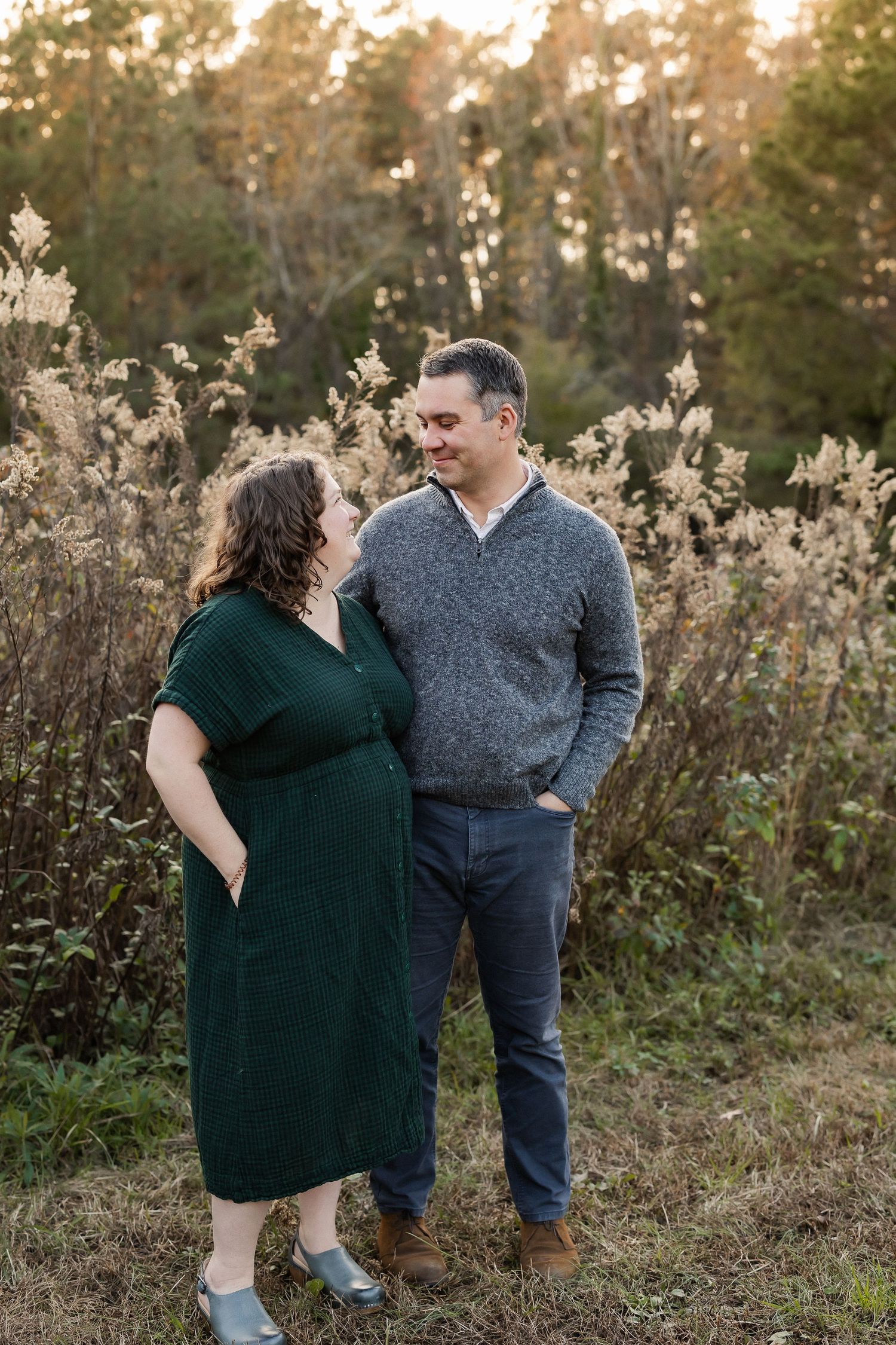 A couple stands together in a field of tall grass during golden hour, wearing casual fall attire.