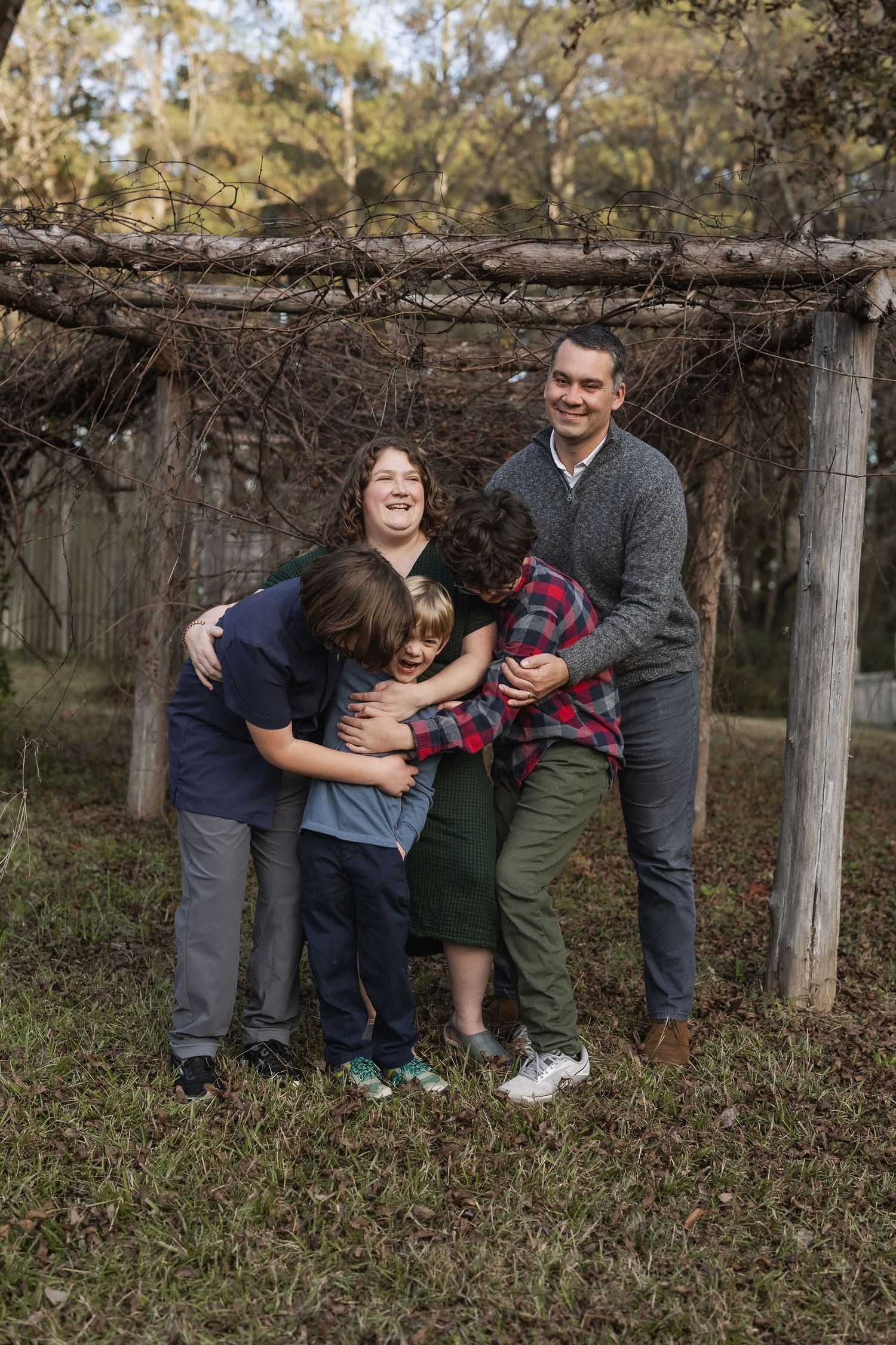 A family stands together embracing under a rustic wooden pergola in an outdoor woodland setting.