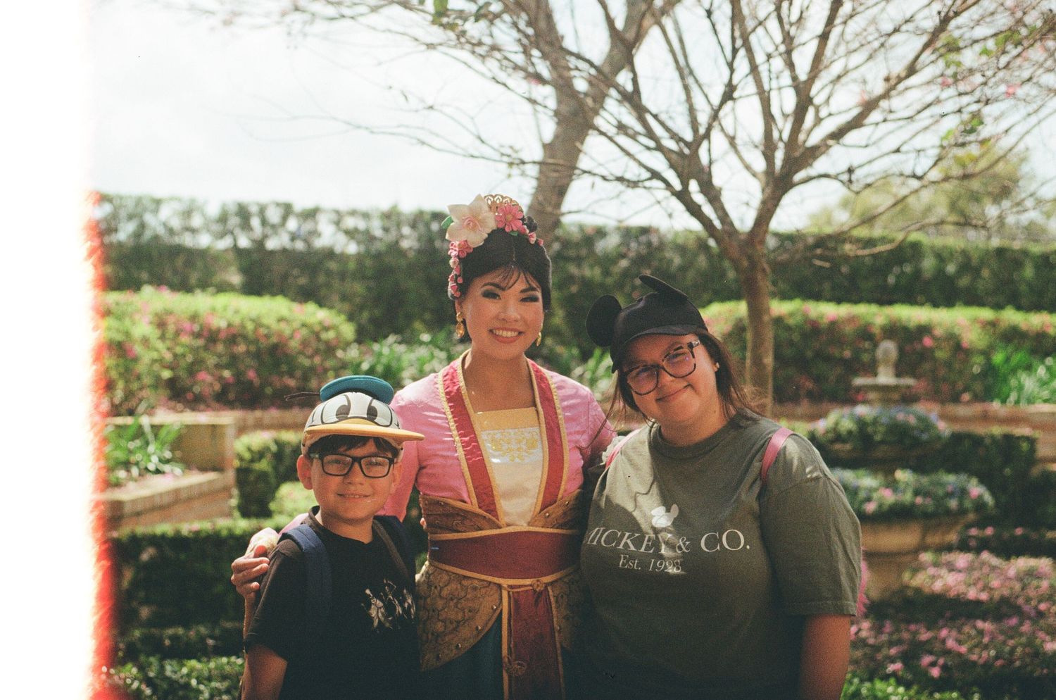 Theme park visitors posing with costumed character in garden setting.