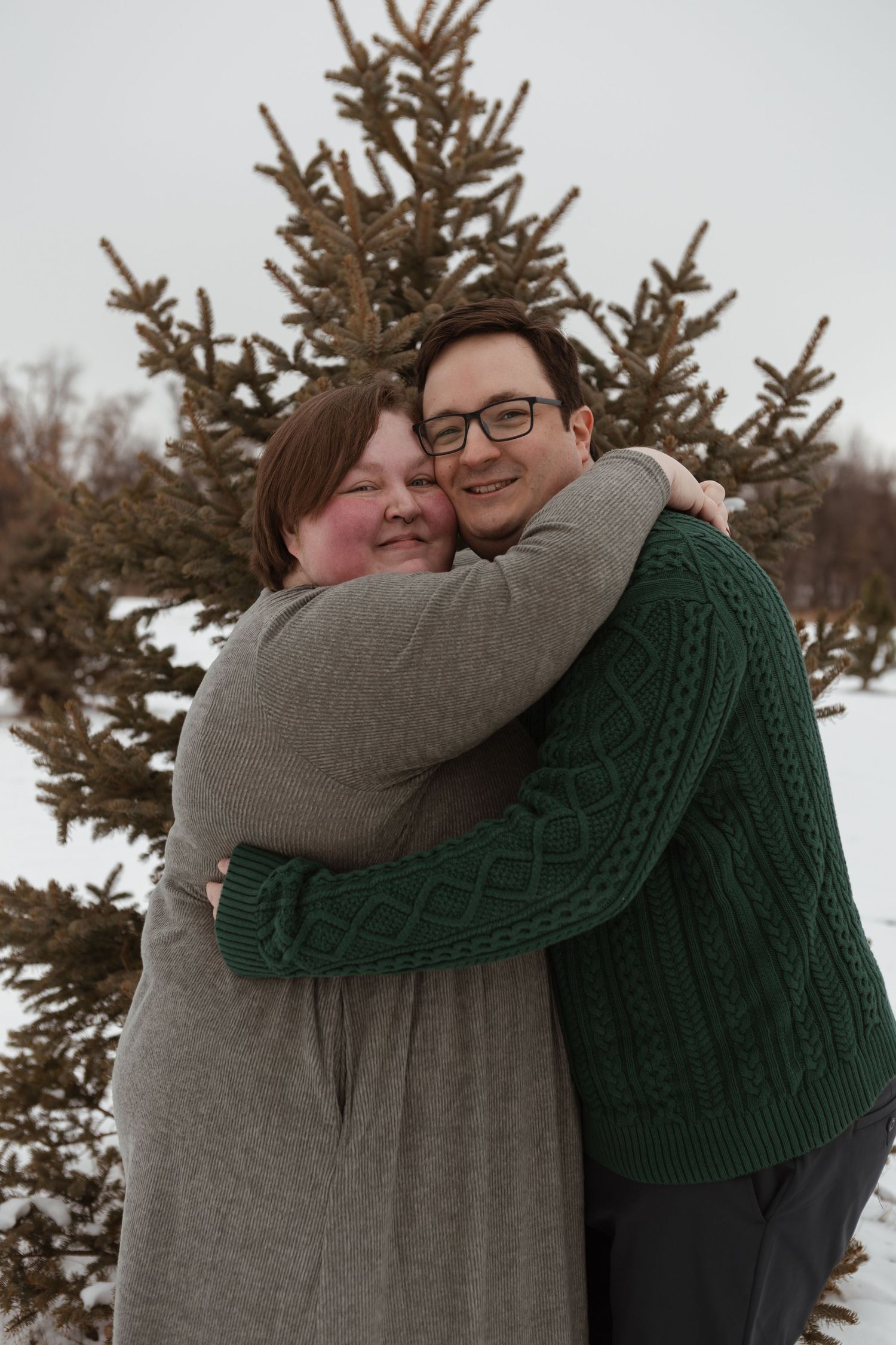 A couple in cozy winter sweaters embrace in front of an evergreen tree on a snowy day.