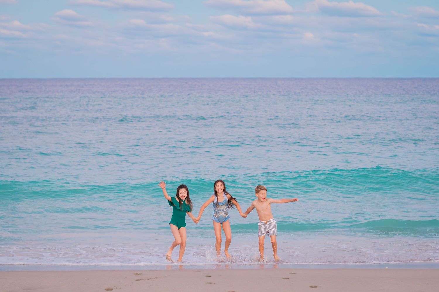 Three children hold hands while joyfully running together along a tropical beach with turquoise waters and pink sky.