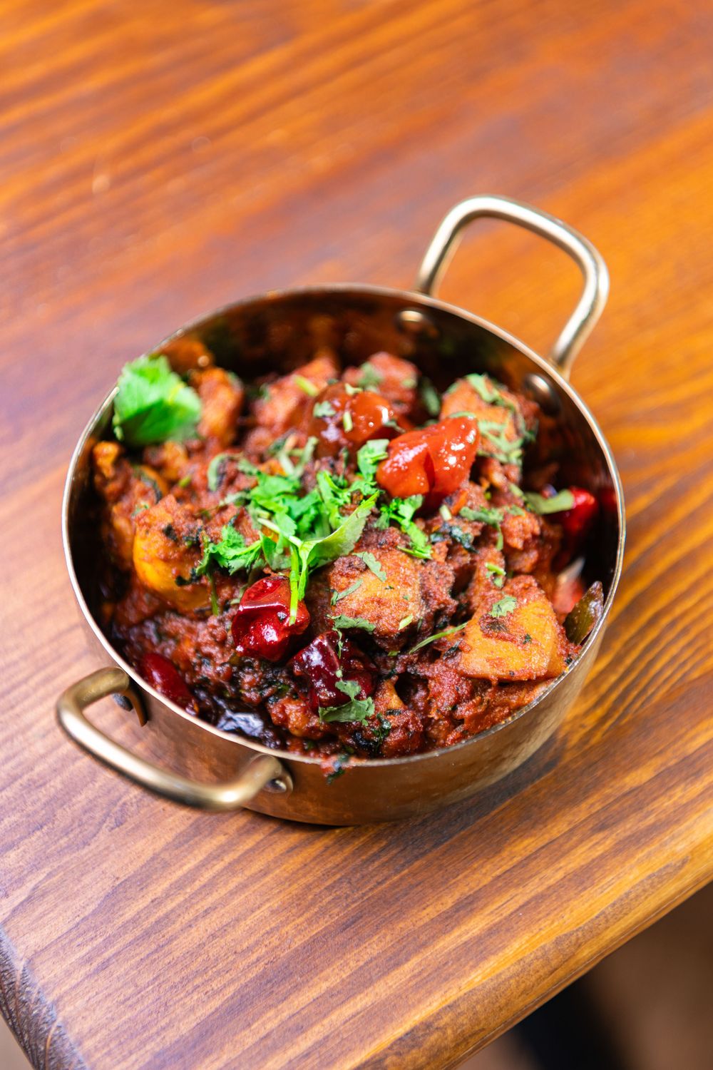 Spicy Indian curry served in a traditional brass kadai bowl, garnished with fresh cilantro and red chilies on a wooden table.
