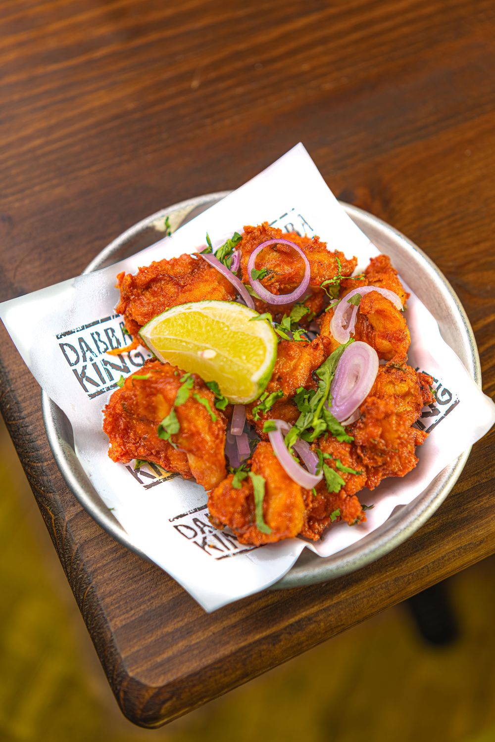 Crispy fried chicken pieces garnished with fresh cilantro, red onion rings, and lime wedge served on paper at Dabba King.