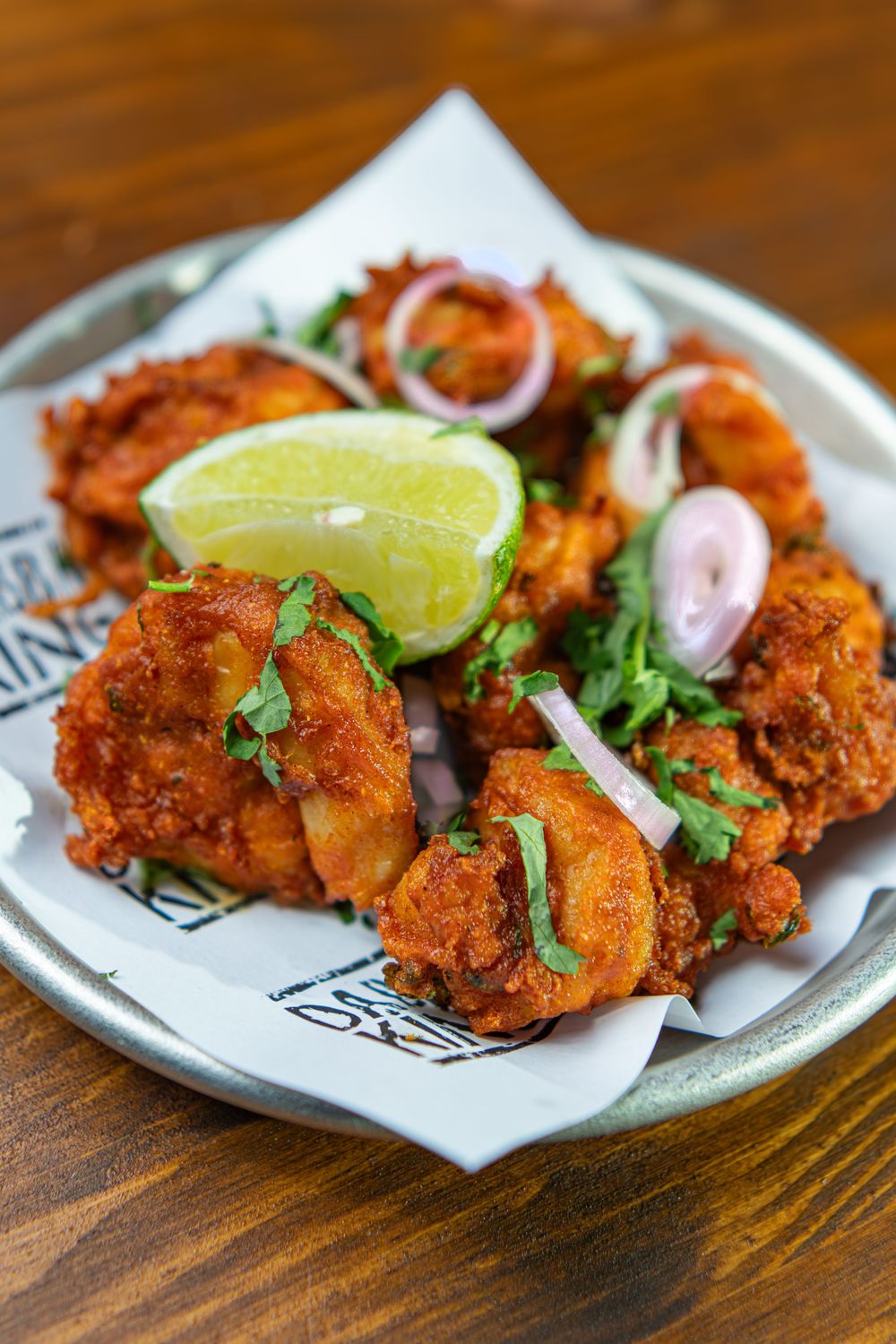 Crispy fried chicken pieces garnished with fresh cilantro, red onion rings, and a lime wedge on a silver plate.