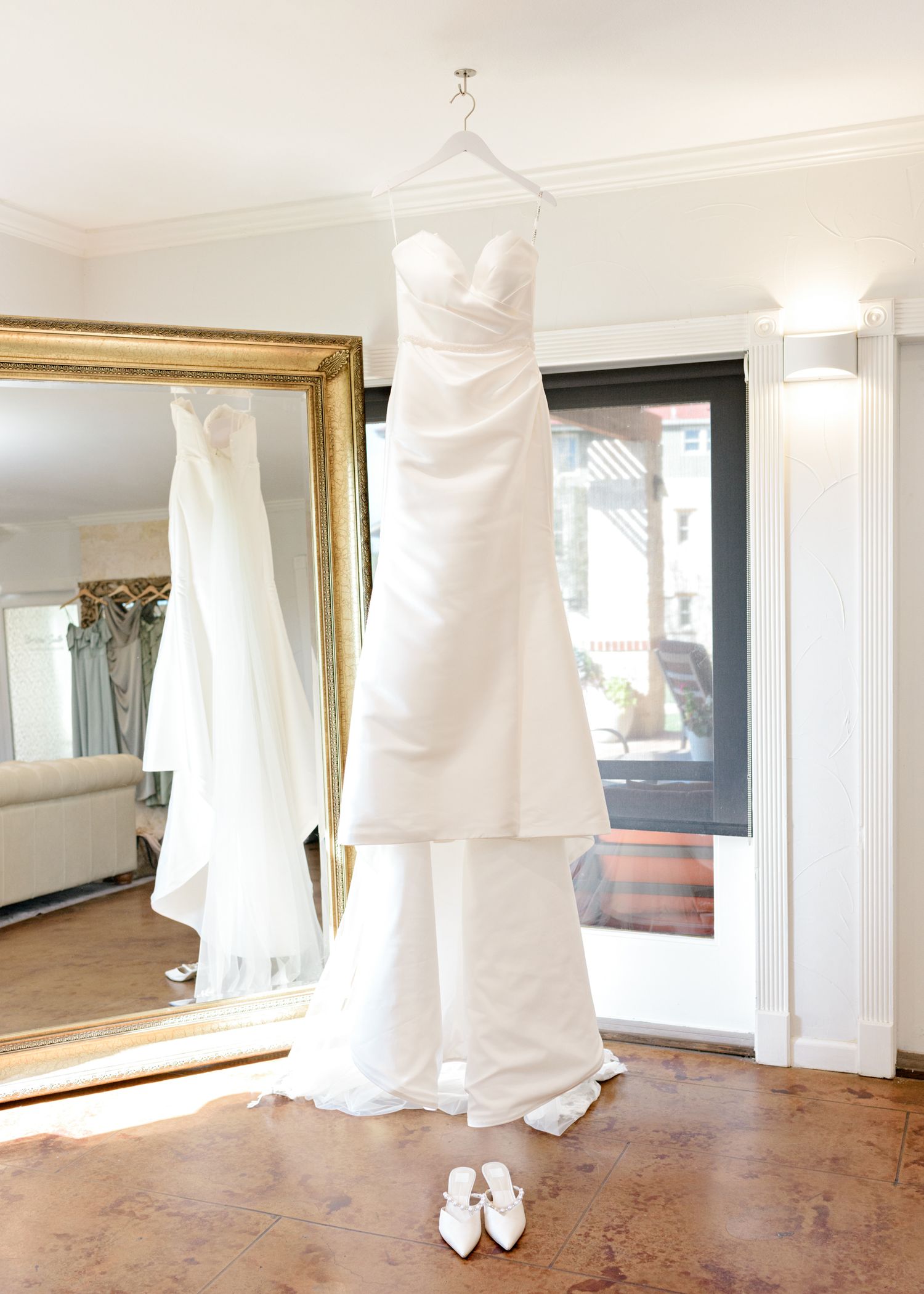 Elegant white wedding gown displayed on hanger in bright dressing room with gold-framed mirror.
