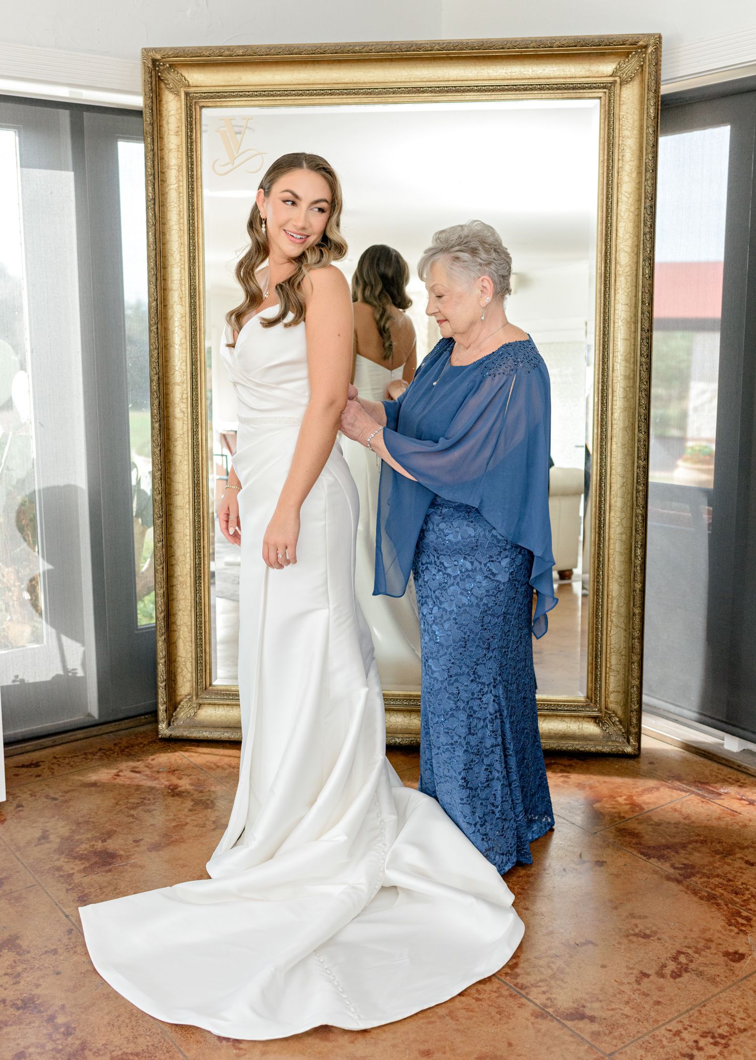A wedding dress fitting scene with a long white gown and elegant blue dress in front of a gold mirror.