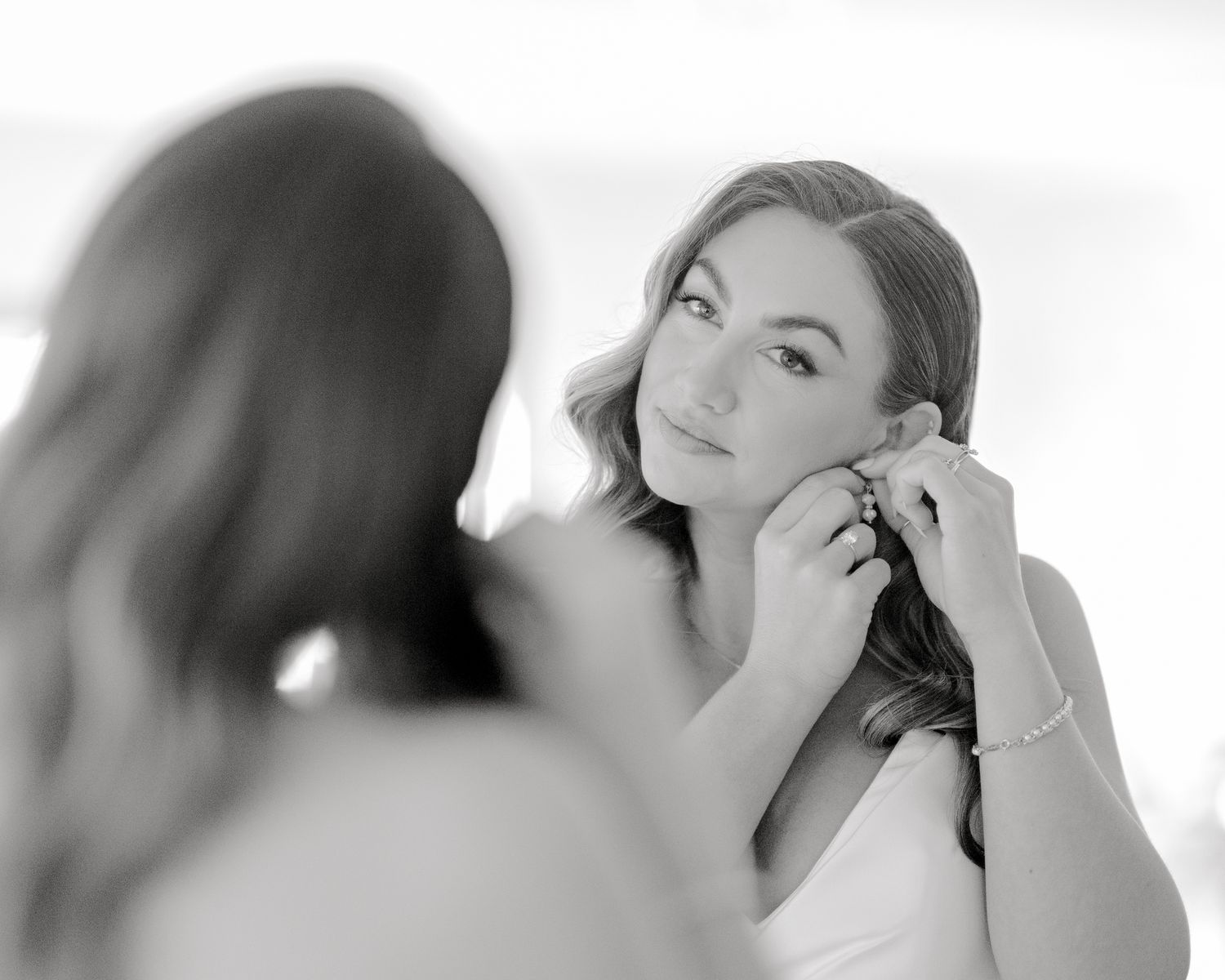 A black and white close-up of someone putting on earrings while looking in a mirror.