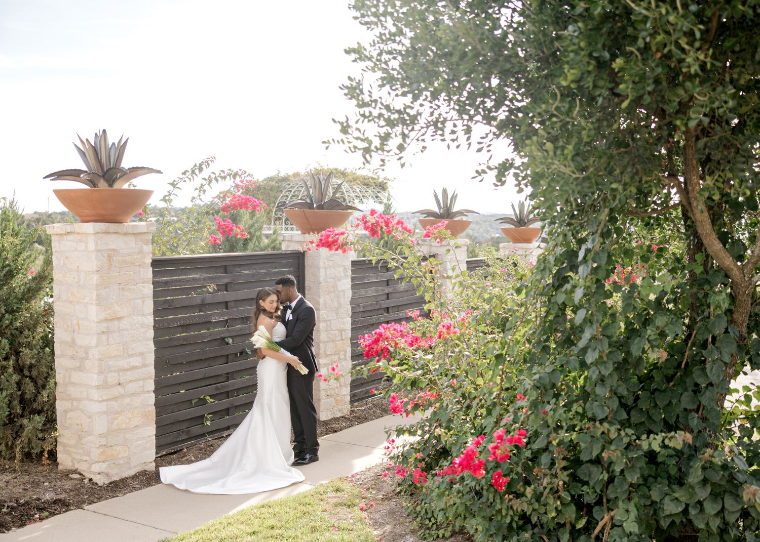 Wedding couple poses in an elegant garden setting with stone pillars and pink flowering vines.