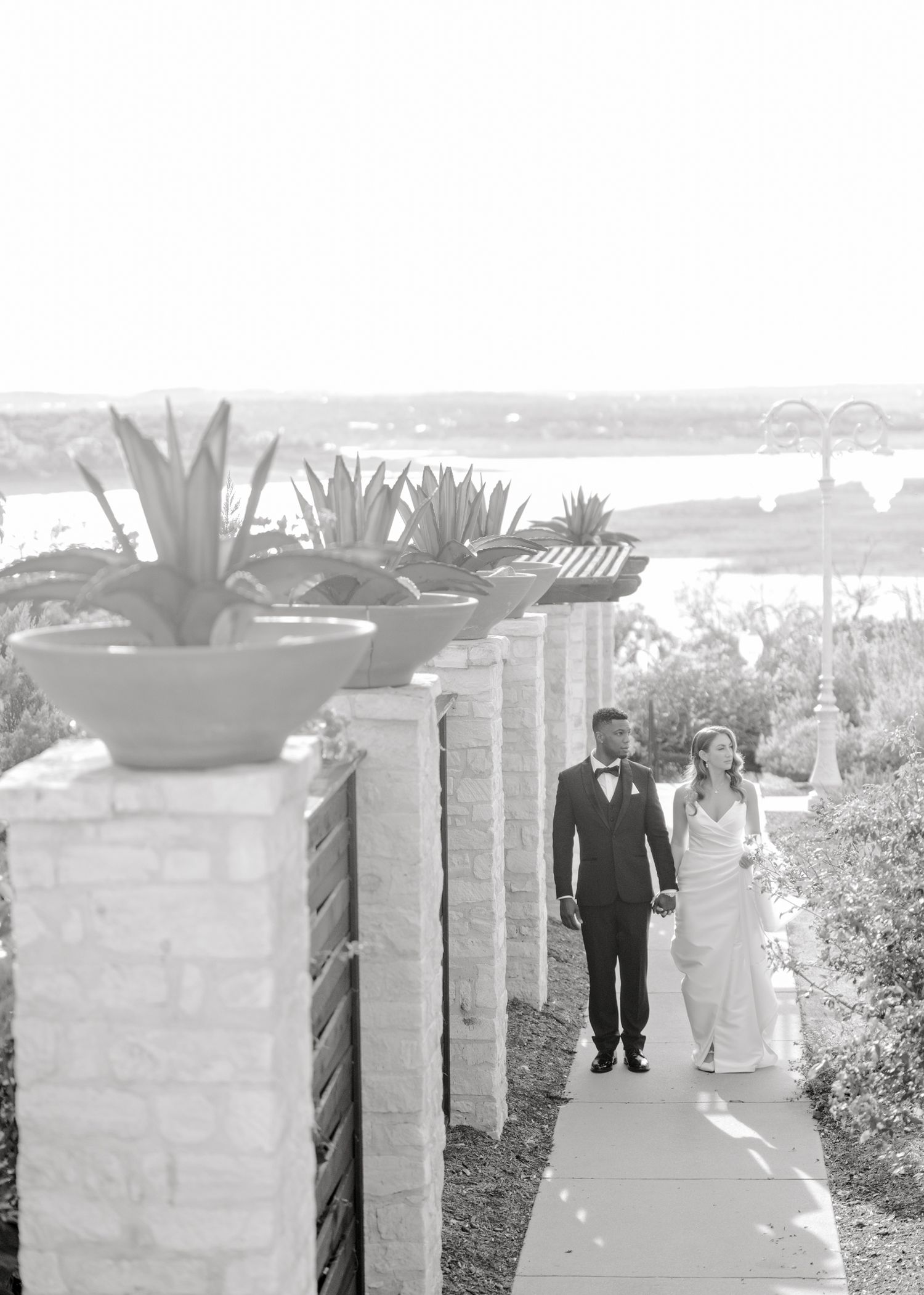 Elegant black and white photo of newlyweds walking along a waterfront pathway.