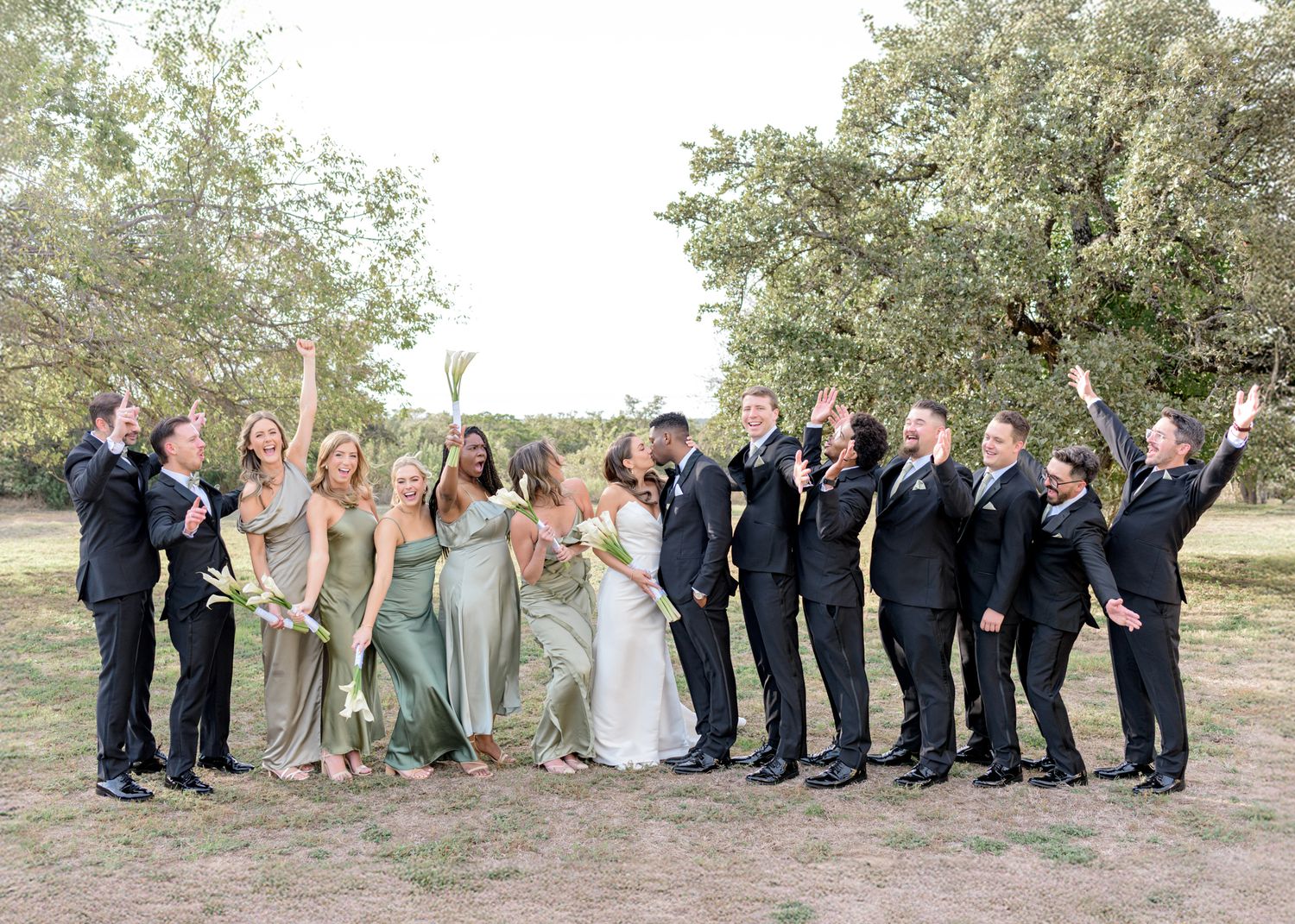 Wedding party poses joyfully outdoors in formal attire against natural backdrop.