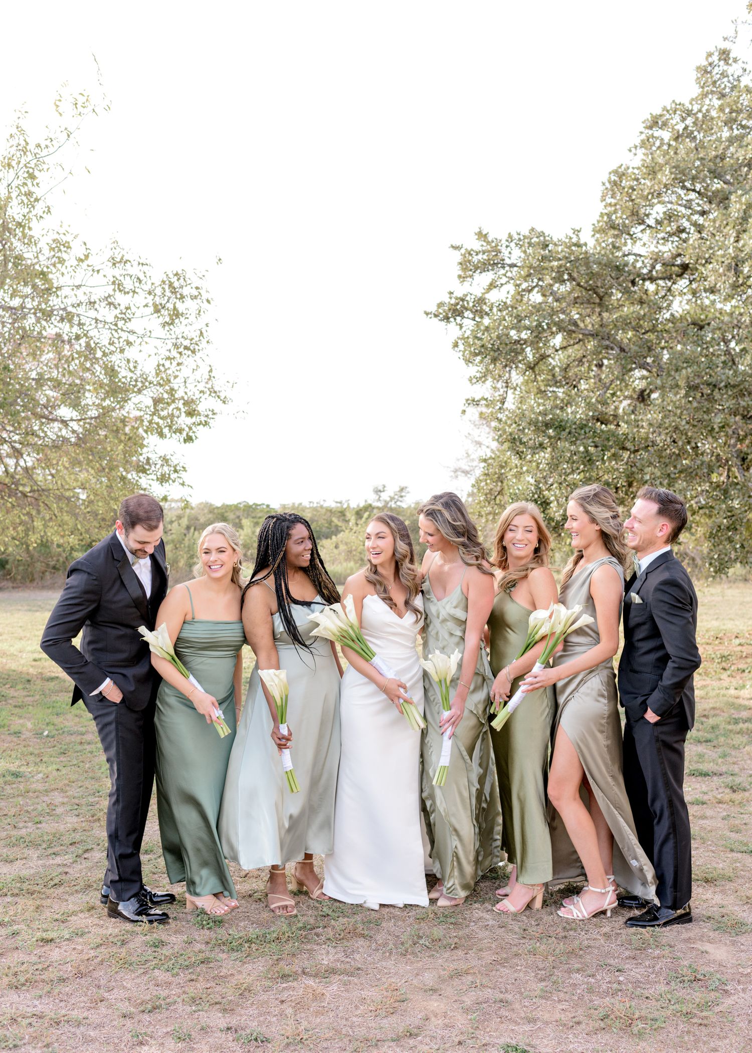 Bridal party in sage green dresses and black suits pose together outdoors.