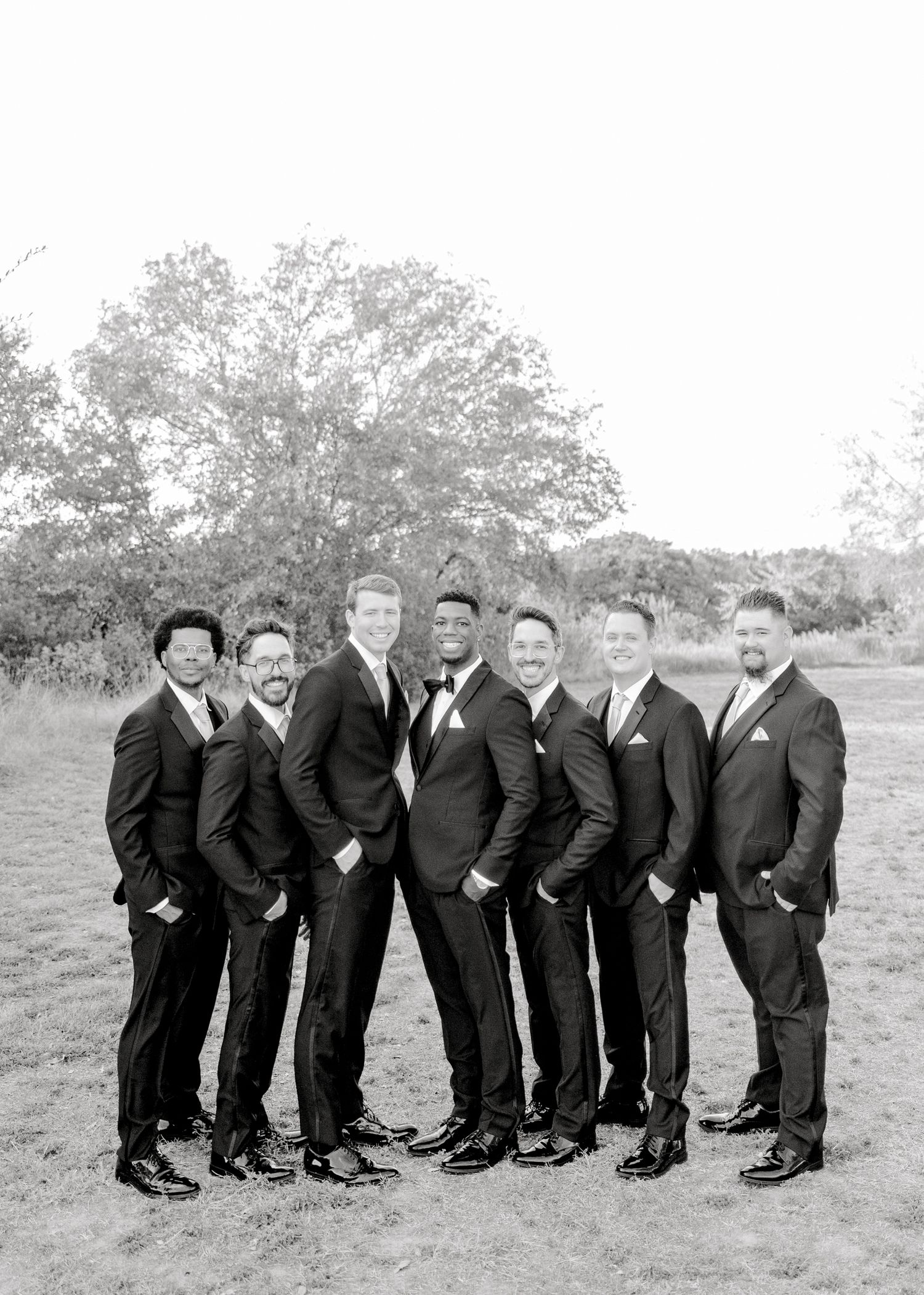 Groomsmen in dark suits pose together for formal portrait outdoors.