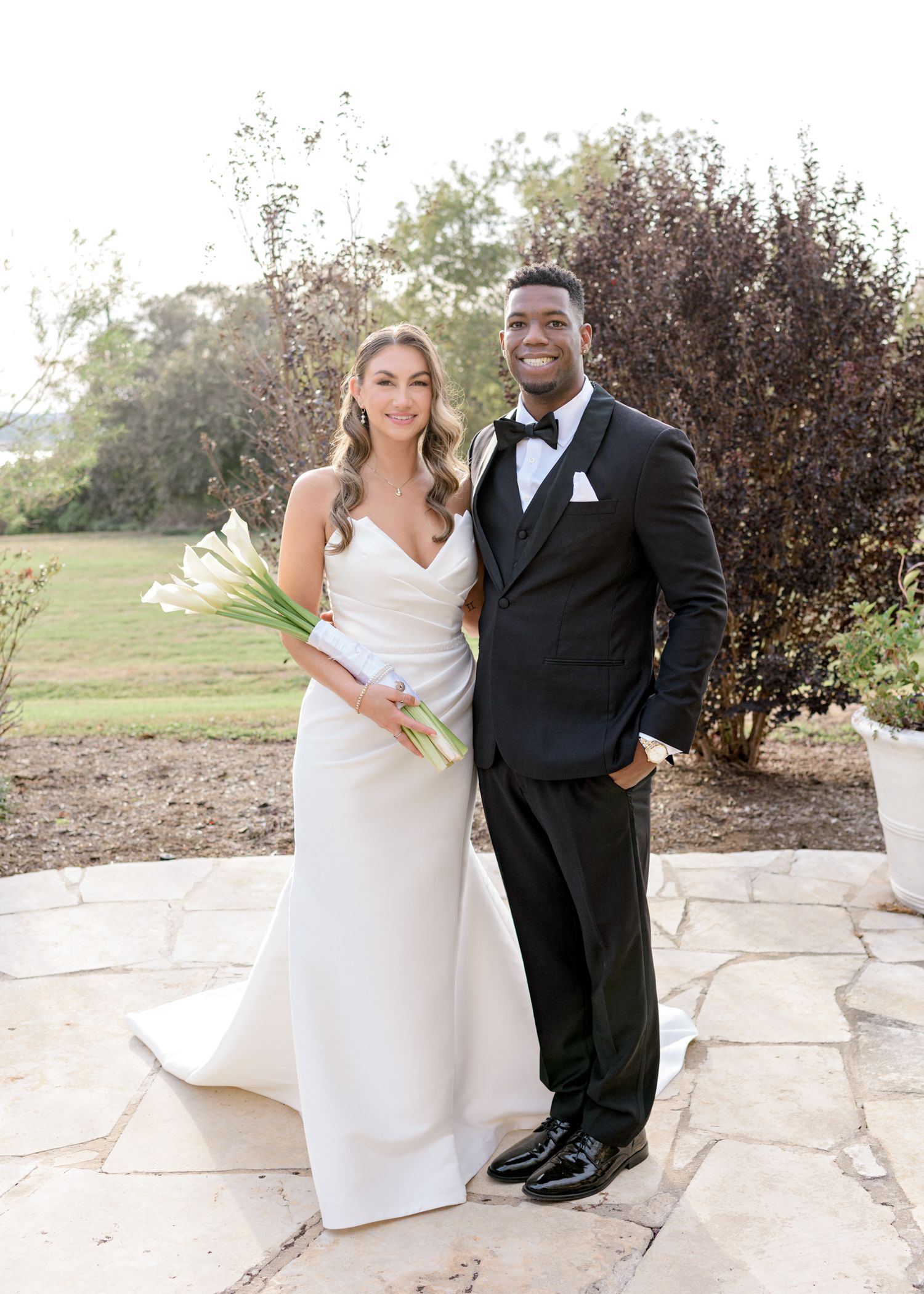Elegant bride in white dress holds calla lilies while posing with groom in classic black tuxedo on stone patio.