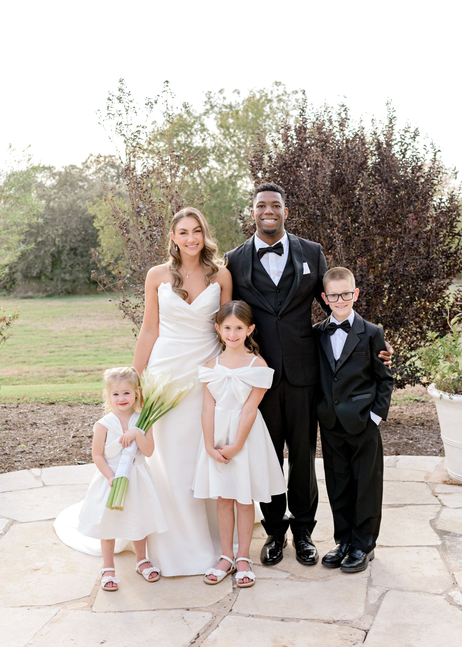Wedding party portrait with bride and groom in formal attire alongside two children in matching outfits on stone patio.
